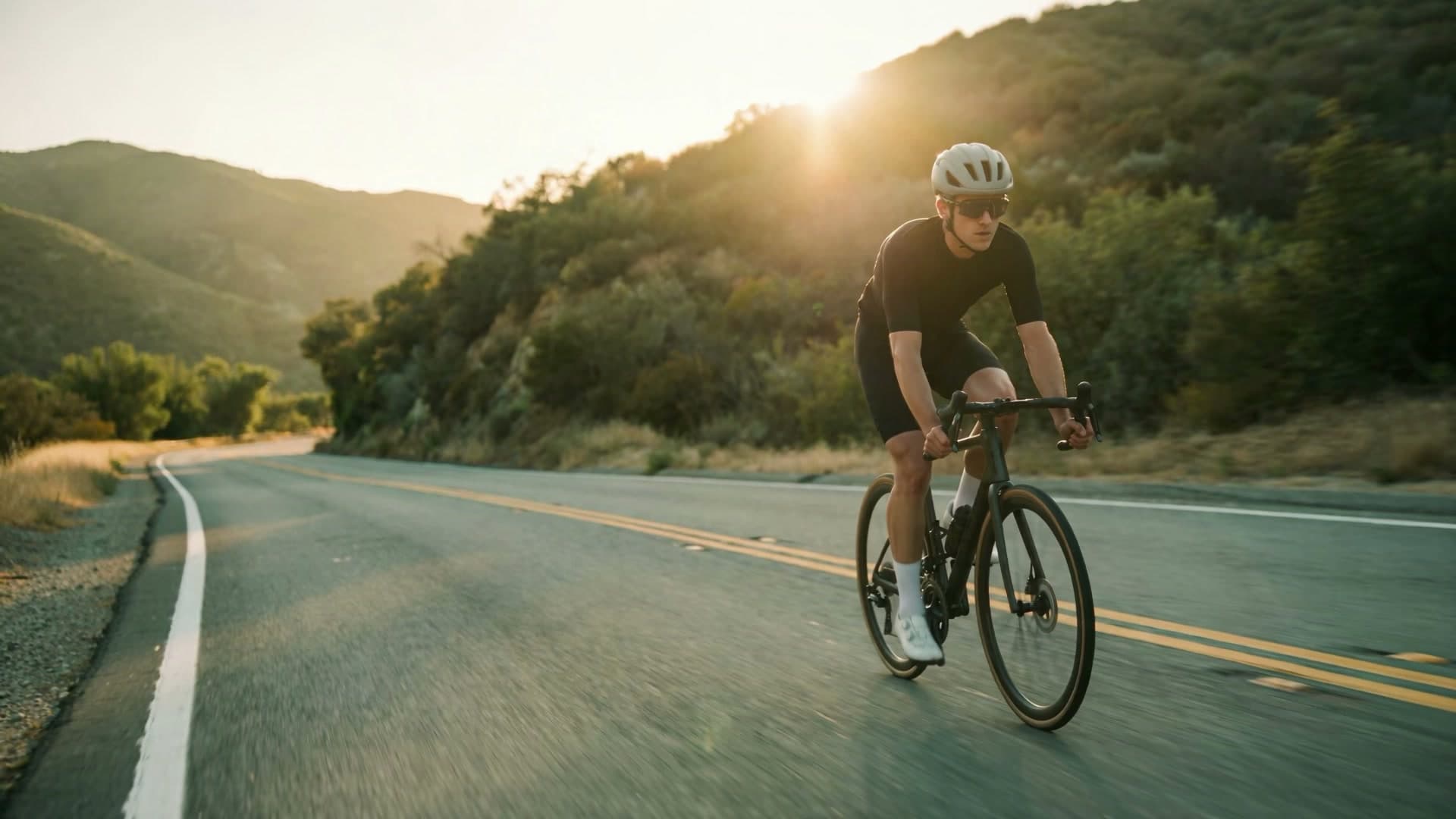 A cyclist descends a winding mountain road towards the camera, backlit by the low evening sun through the trees.