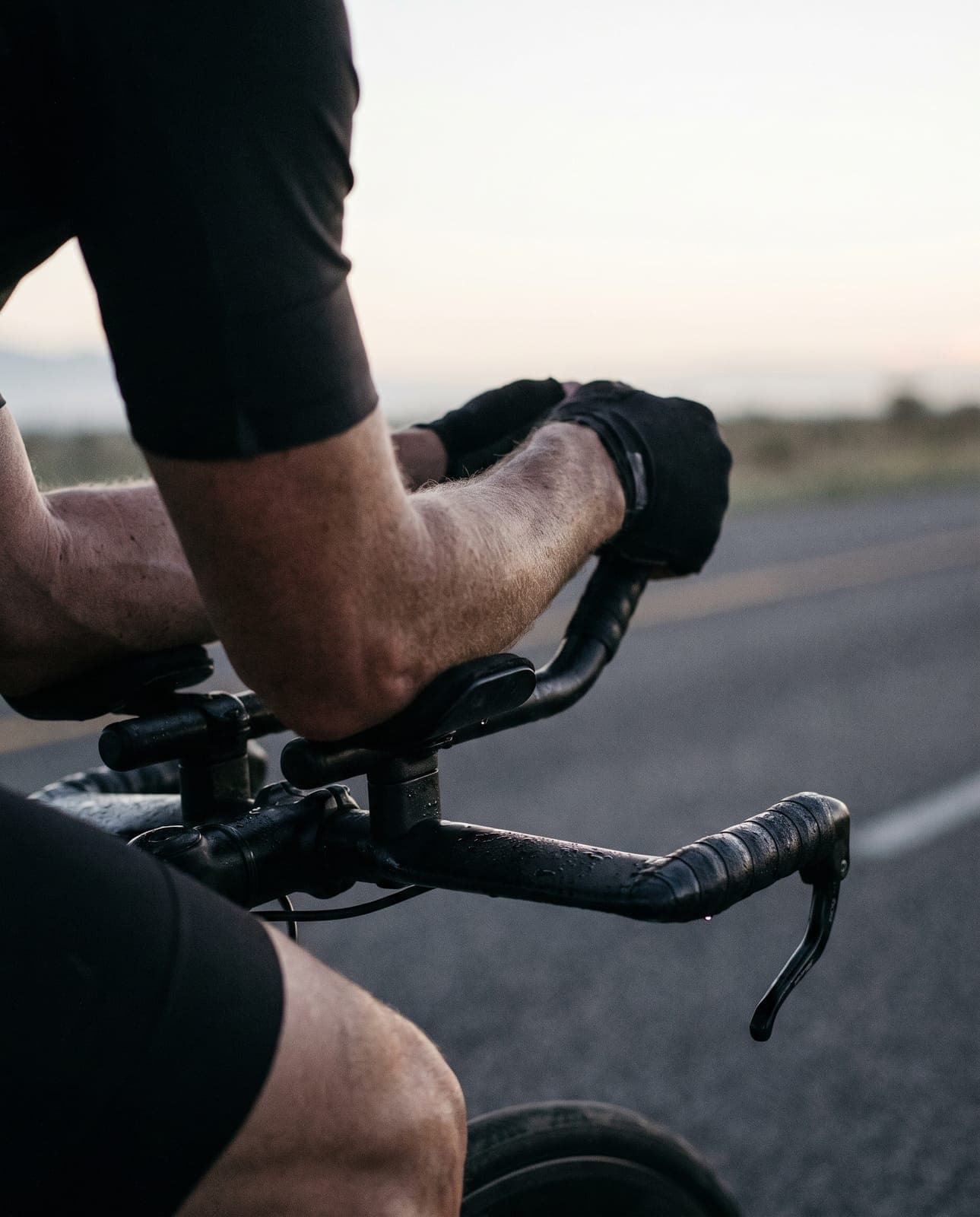 Antebrazos y manos apoyados en las extensiones de una bicicleta de contrarreloj al amanecer, con la carretera desenfocada bajo la primera luz.